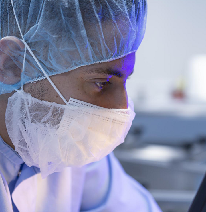 A scientist wearing a facemask and a headcap looking into a monitor